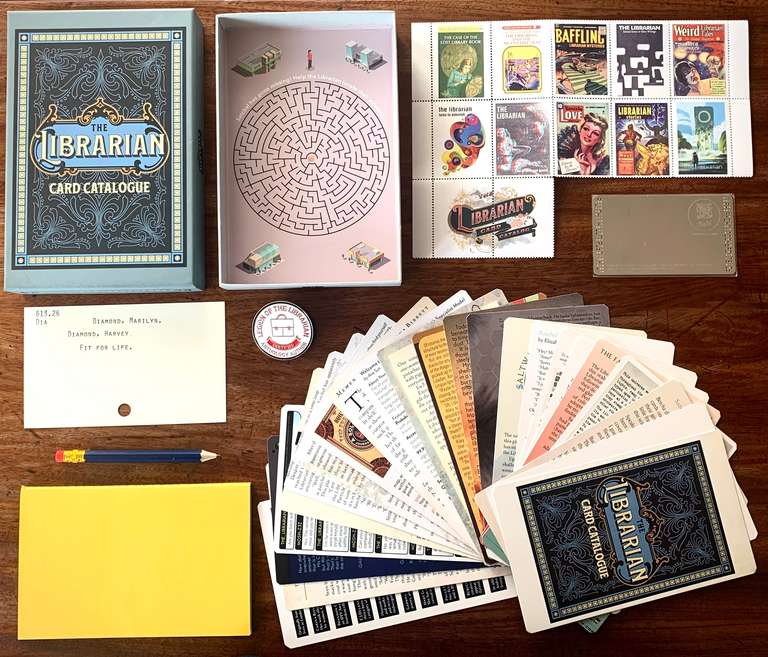 Catalalogue contents include: 23 oversized playing cards, a metal library card, a saddle stitched notebook and pencil, and 12 postage stamps in a rigid box